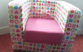 A small, boxy armchair for children covered in colourful patterns of hearts, flowers, and fruit shapes. The seat cushion is solid pink. The chair sits on a red carpet in front of a white wall with a narrow skirting board.