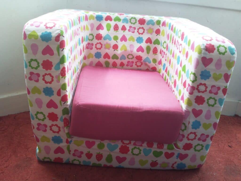 A small armchair for children with a bright, multicoloured pattern of hearts, flowers, and strawberries covering the sides and back. The seat cushion is solid pink. The chair sits on a red carpet next to a white wall and skirting board.