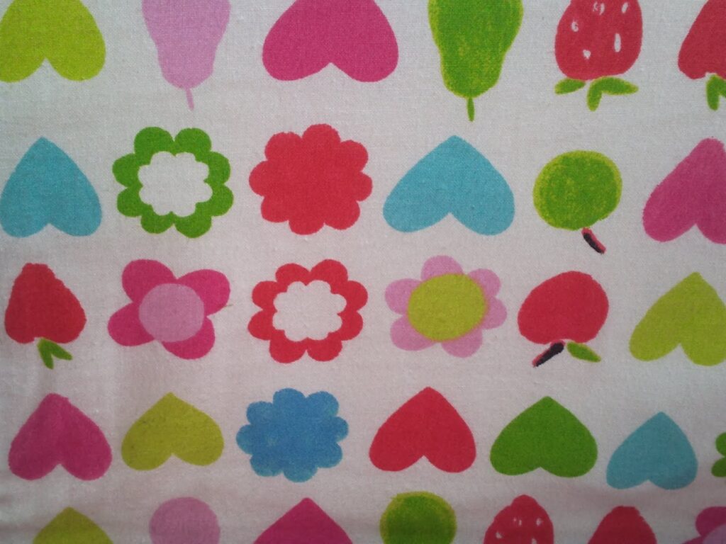 A colourful pattern on fabric featuring hearts, flowers, strawberries, and apples in shades of pink, red, blue, green, and white, arranged in a grid on a white background. The design is playful and vibrant.