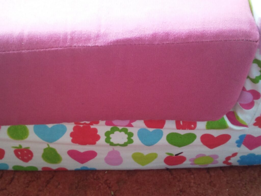A close-up of a piece of furniture features a pink fabric cushion on top and a base covered in white fabric with colourful prints of hearts, flowers, strawberries, and apples in shades of blue, red, pink, and green.