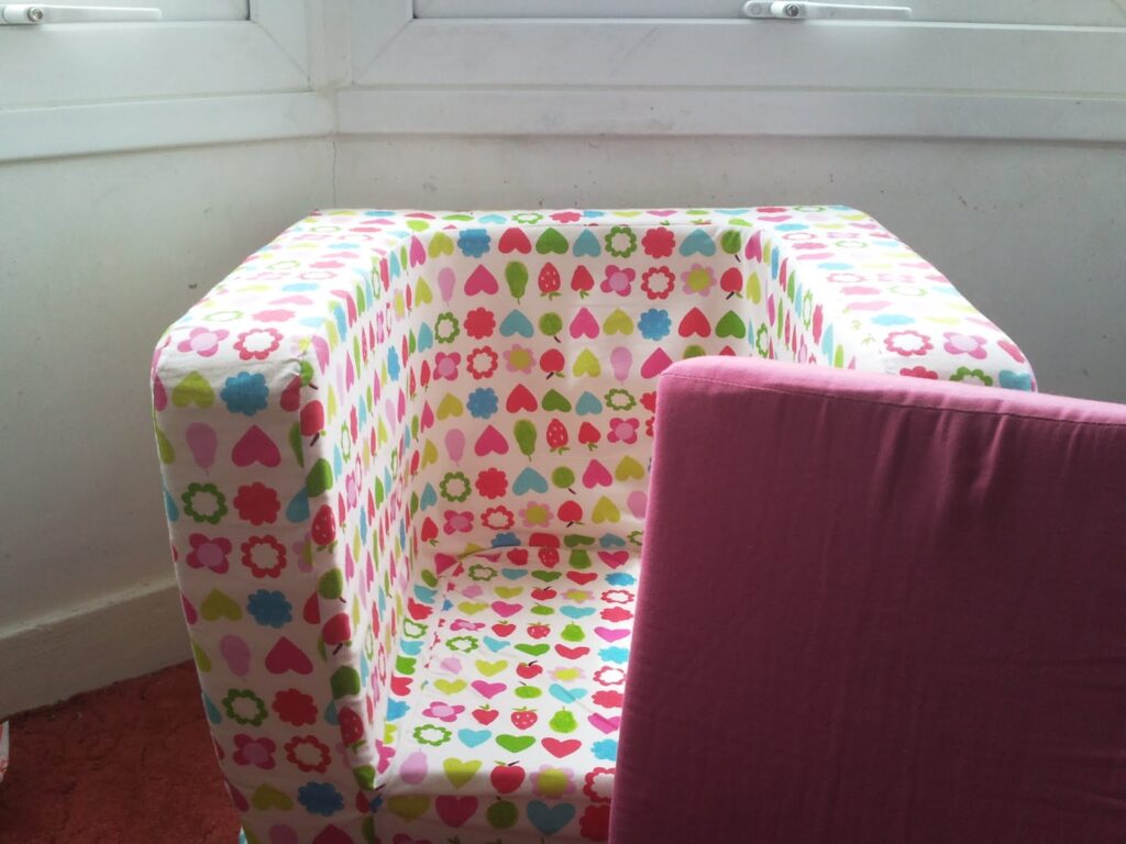 A small armchair covered in fabric with a colourful pattern of hearts, flowers, and shapes sits in a corner by white-painted windows. A plain pink cushion or panel leans against the chair. The floor is carpeted in a reddish shade.