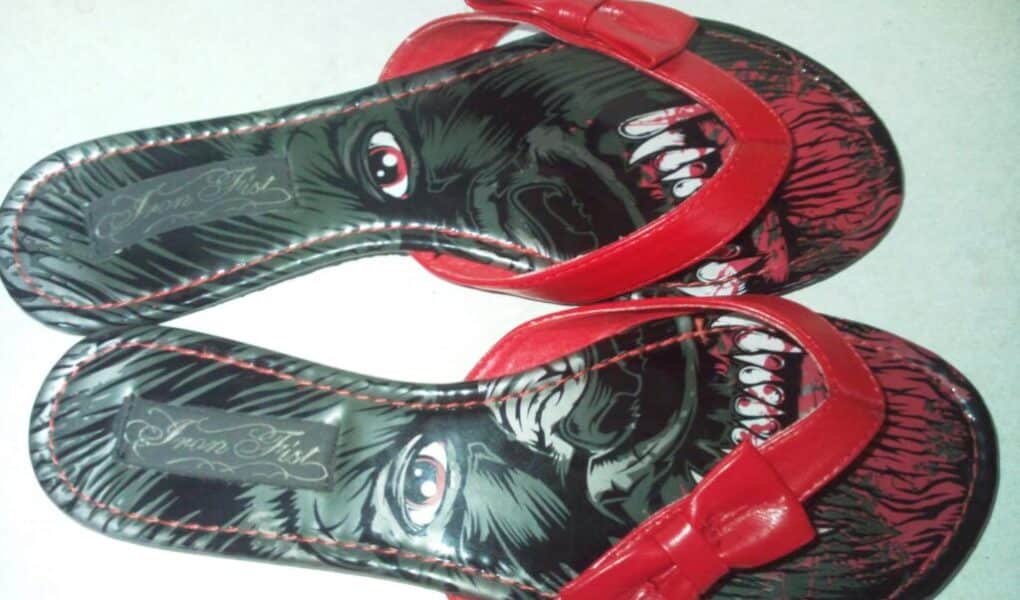 A pair of red sandals with a unique design; the insoles feature an illustration of a face split across both shoes—one sandal shows an intense eye, the other a snarling mouth with teeth. The face combines when the sandals are placed side by side.