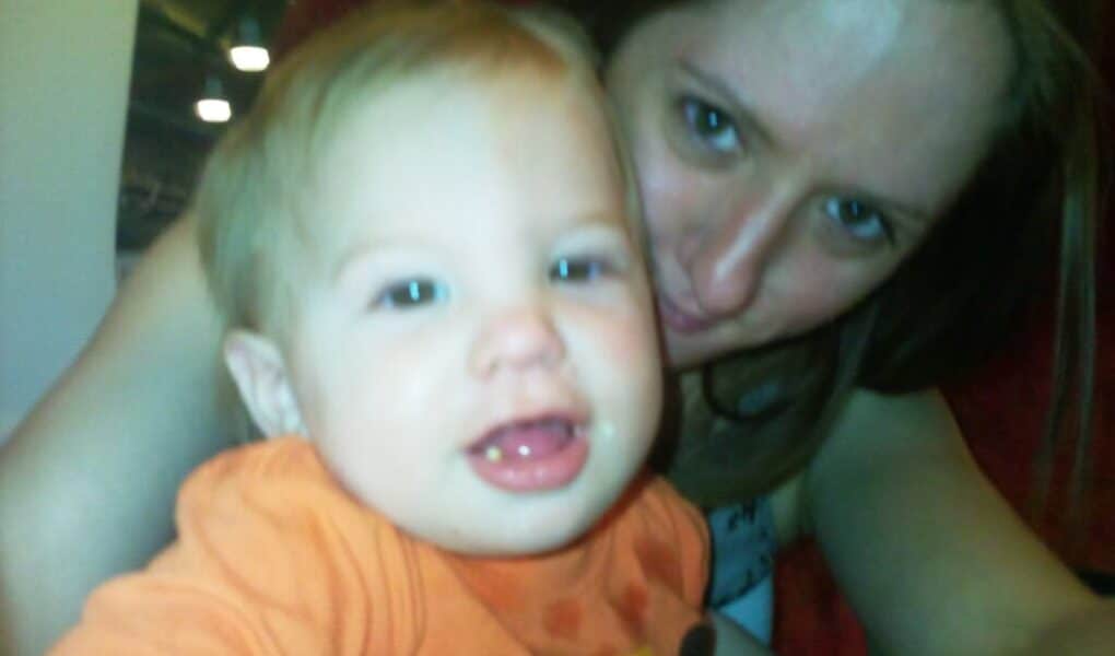 A woman with light brown hair is smiling closely behind a happy baby wearing an orange shirt with a cartoon design. The baby is looking at the camera with mouth open, showing a tooth. The image is brightly lit and slightly blurred.