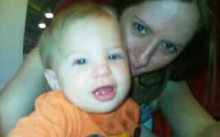 A woman with light brown hair is smiling closely behind a happy baby wearing an orange shirt with a cartoon design. The baby is looking at the camera with mouth open, showing a tooth. The image is brightly lit and slightly blurred.