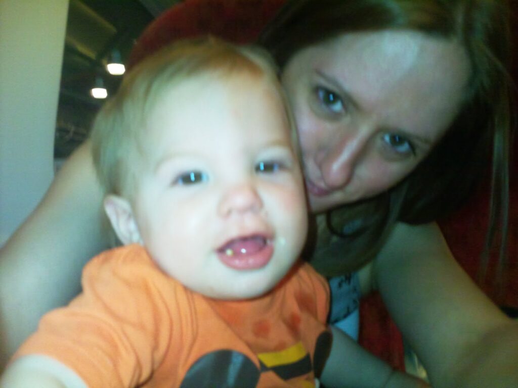 A smiling baby in an orange shirt sits close to a woman with light brown hair, who is smiling and leaning towards the child. The photo is slightly out of focus, giving a warm and candid feeling to the moment.