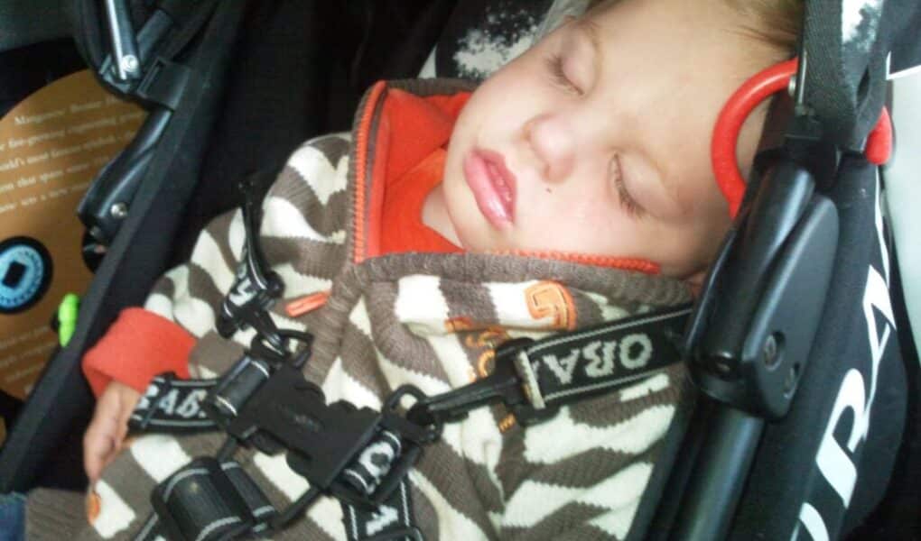 A young child is sleeping peacefully in a pram, secured with a harness. The child wears a brown and white striped jacket with orange accents and rests their head against the pram’s side. The child’s eyes are closed and mouth slightly open.