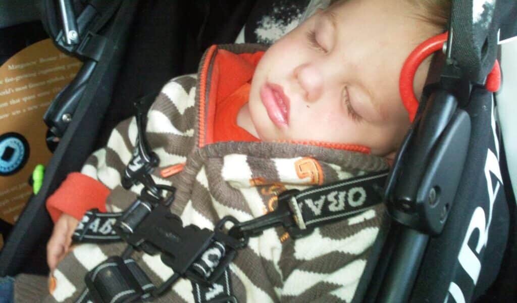 A young child is sleeping peacefully in a pram, secured with a harness. The child wears a brown and white striped jacket with orange accents and rests their head against the pram’s side. The child’s eyes are closed and mouth slightly open.