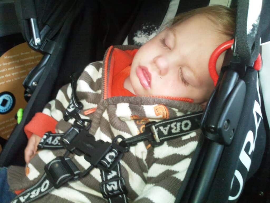 A young child with light skin and short hair sleeps peacefully in a pushchair, wearing a brown and white striped jumper with an orange shirt underneath. The pushchair harness is fastened securely across the child's chest. The child’s head is tilted to the side.