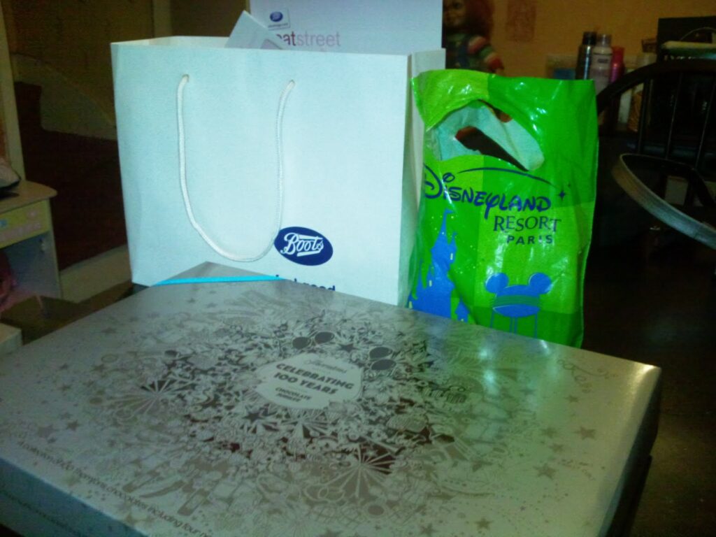 A silver box with decorative designs sits on a table in front of a white Boots shopping bag and a green Disneyland Paris bag. The background includes a black chair and various household items in a dimly lit room.