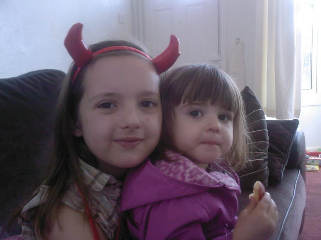 Two young girls sit on a dark settee. The older girl wears a red headband with devil horns and smiles softly, hugging the younger girl, who has straight brown hair and wears a pink hoodie while holding a snack. Light enters through a window behind them.