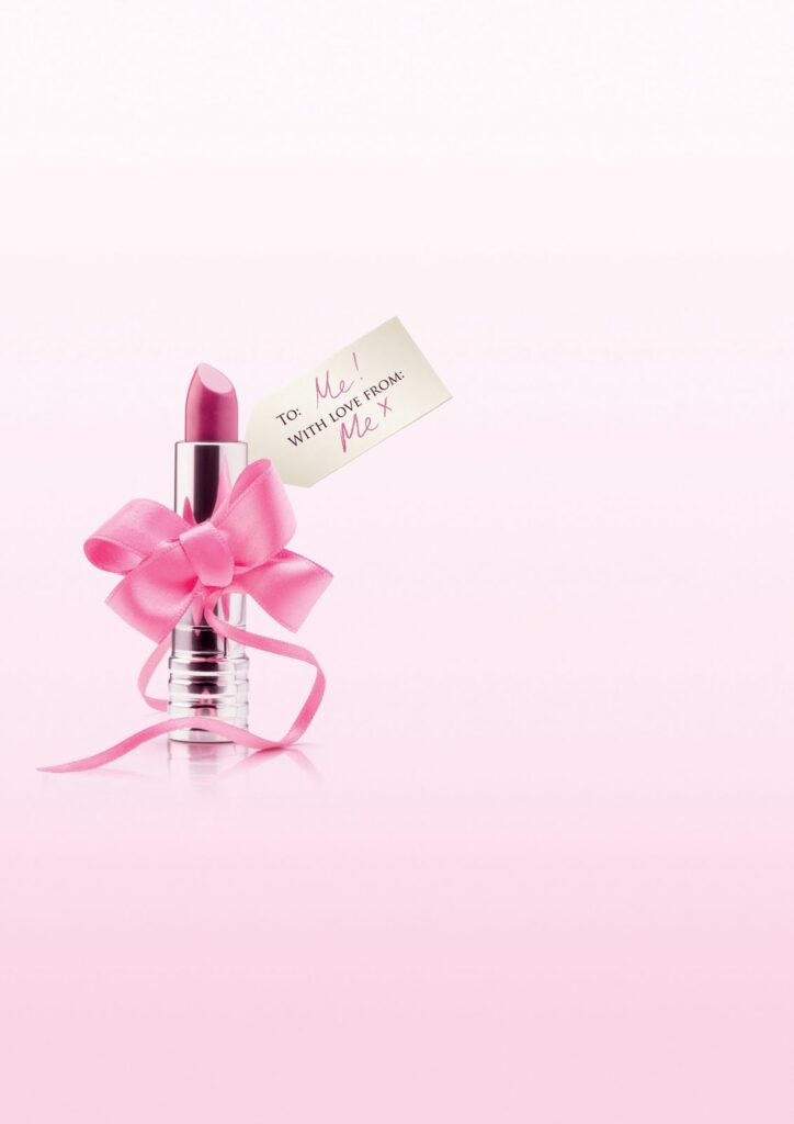 A pink lipstick in a silver tube is wrapped with a pink ribbon bow. Attached to the ribbon is a tag that reads, "To: Me! With love from: Me x." The background is a soft pink gradient fading to white at the top, creating a light and cheerful mood.