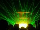 A crowd of silhouetted people stands before a stage filled with bright green laser lights fanning out in various directions. The intense yellow and green glow at the centre suggests a concert or music event with dramatic lighting effects.