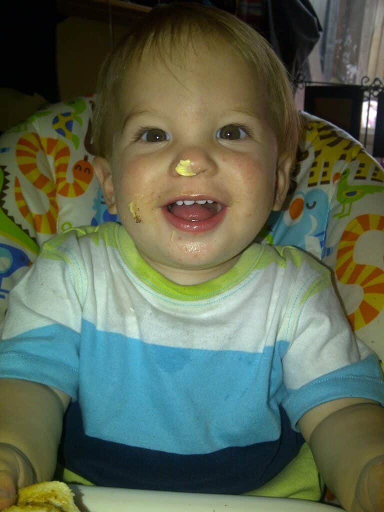 A smiling toddler with fair hair sits in a colourful high chair. The child has a dollop of food on their nose and some on their cheek, wearing a blue and white shirt, holding food with both hands, and looking up in a cheerful, playful mood.