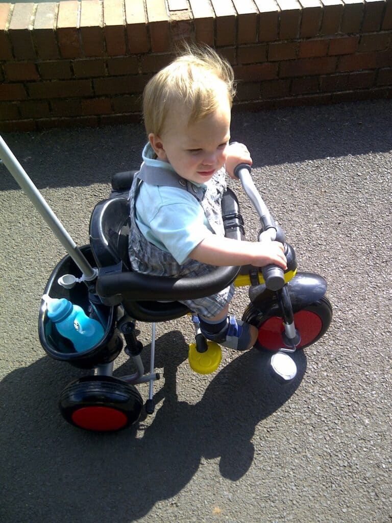 A young toddler with blond hair sits on a black and red tricycle outdoors, gripping the handlebars. The tricycle has a blue bottle in the back basket, and sunlight casts shadows on the paved ground. A brick wall is in the background.