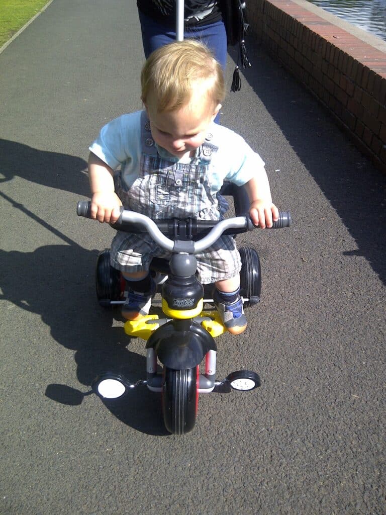 A young toddler with blond hair wearing a blue shirt and checked dungarees rides a black and yellow tricycle on a sunny path. An adult is pushing the tricycle using a white handle. Part of a brick wall and water are visible to the right.