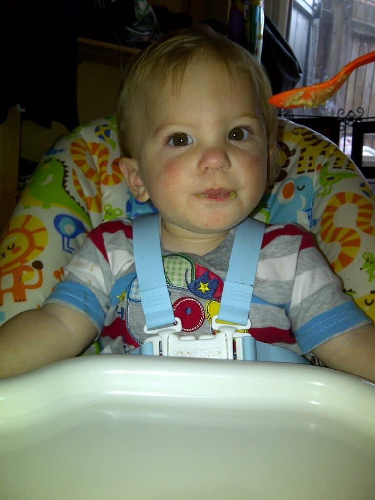 A young child with fair hair sits in a highchair, wearing a striped shirt and blue harness. The highchair has a colourful animal pattern. The child looks slightly upward with a neutral expression, resting both hands on the tray in front.