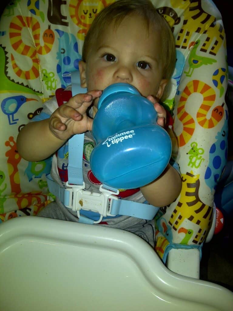 A baby with light brown hair sits in a colourful animal-patterned highchair, holding a blue Tommee Tippee sippy cup with both hands, drinking from it. The baby is strapped in with a blue harness and looks directly at the camera.