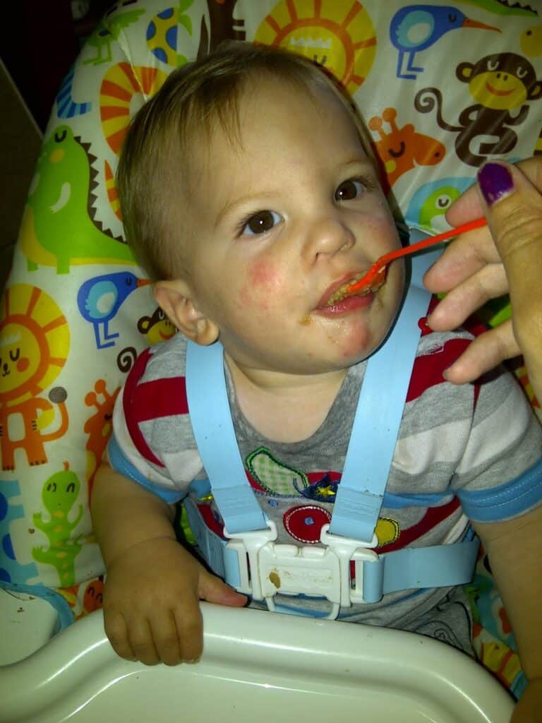 A baby with light brown hair sits in a colourful animal-themed high chair, wearing a striped shirt and blue harness. The baby is being fed with an orange spoon by an adult hand. The baby has food stains around their mouth and some redness on the cheeks.
