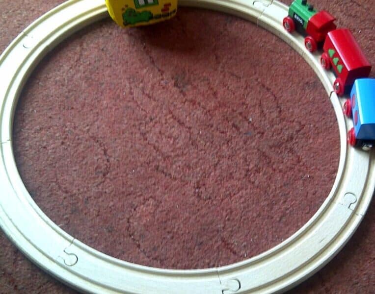 A small toy train with red and blue carriages travels on a circular wooden track set on a reddish carpet. A colourful toy house with a red roof sits partially inside the circle, opposite the train.