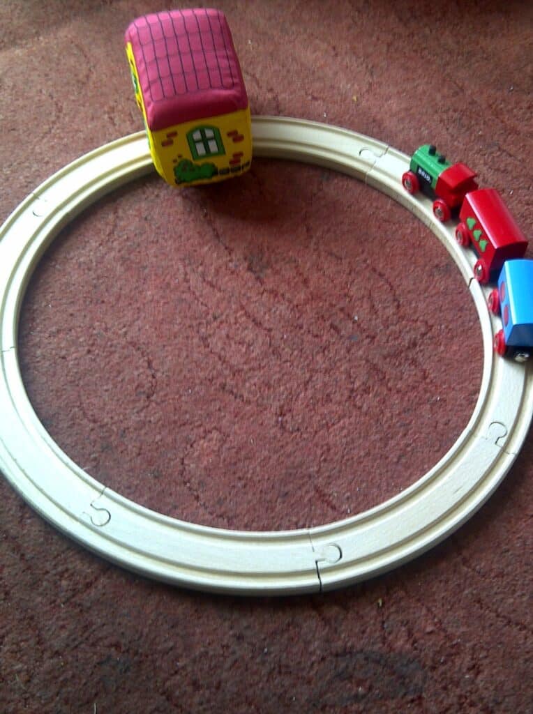 A small wooden toy train with three colourful carriages runs on a circular wooden track. A yellow toy house with a red roof sits inside the track, on a reddish-brown carpeted floor. The train is near the bottom right of the circle.