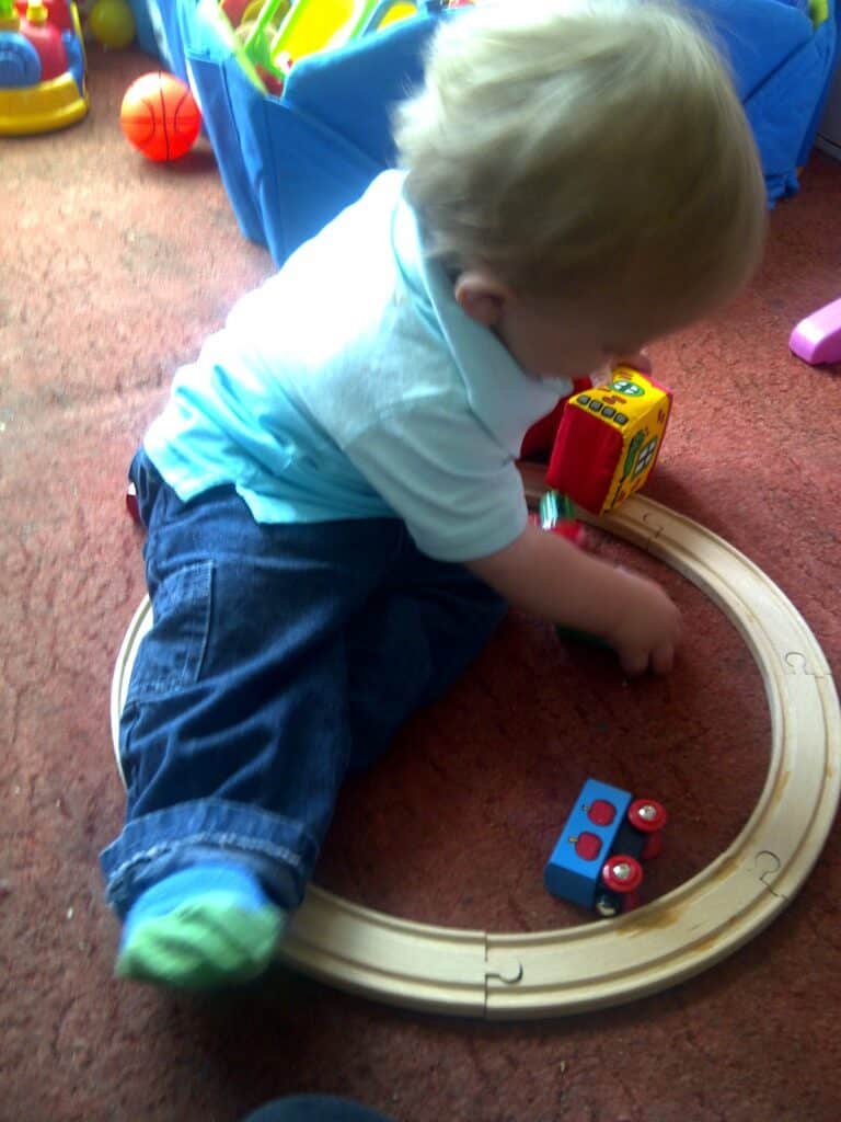A toddler with blond hair sits on a carpet inside a wooden train track. The child wears a light blue shirt, jeans, and green socks, and is playing with colourful toy blocks and a red toy train. Other toys and a blue box are in the background.