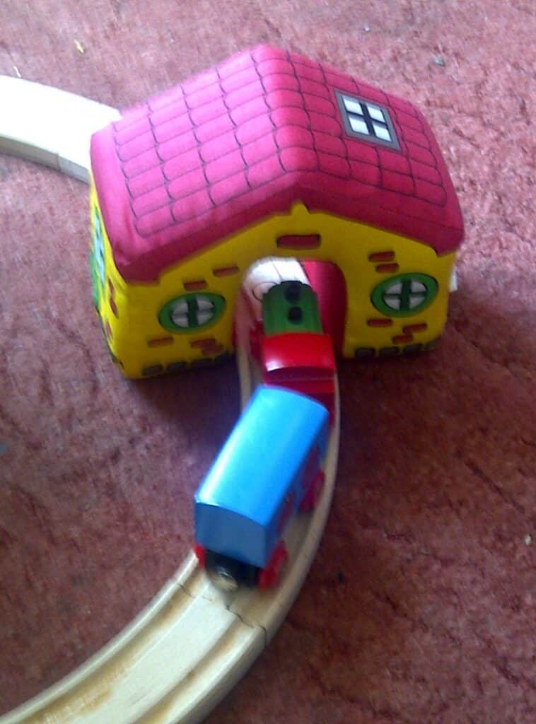 A wooden toy railway track curves through a colourful toy house with red roof and yellow walls. Two toy train carriages, one red and green and the other blue, are on the track emerging from the house, all set on a brown surface.