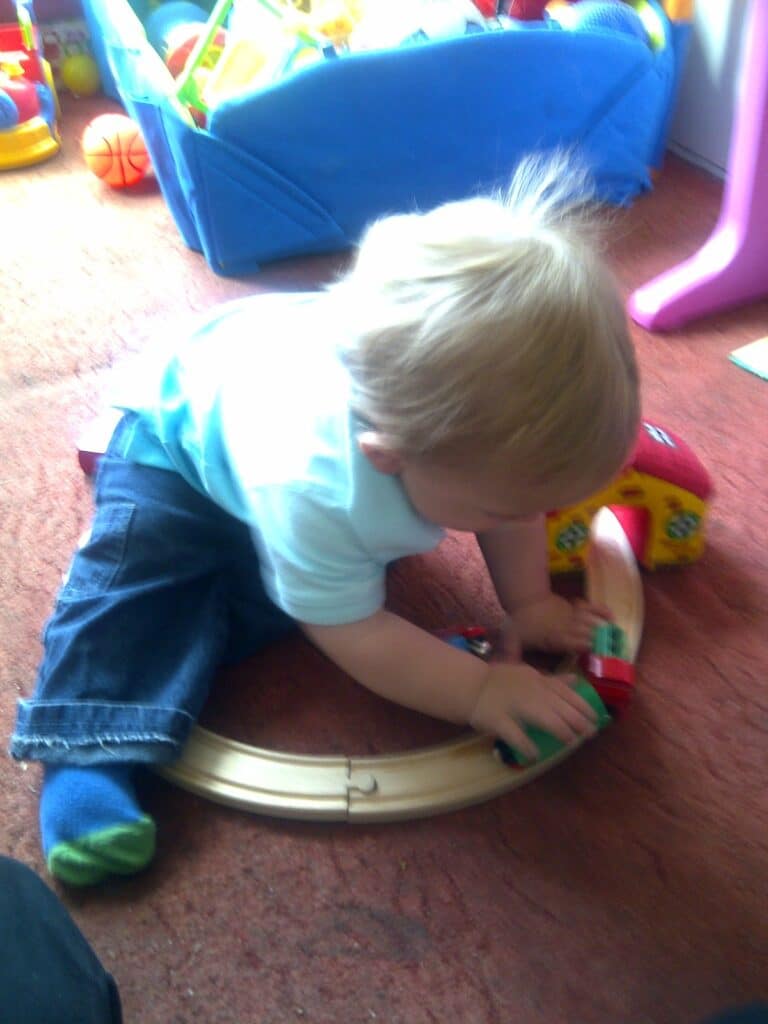 A young child with blonde hair sits on a carpet, playing with a wooden toy train set. The child wears a light blue shirt, dark blue jeans, and colourful socks. In the background, there is a blue toy box filled with various toys.