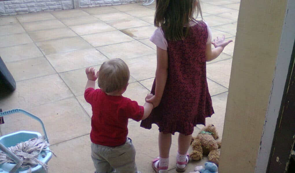 A young girl in a pink and purple dress holds hands with a toddler in a red shirt and light trousers. They stand on a tiled patio near a mop bucket, facing away. Toys, including a teddy bear and a doll, are scattered on the ground beside them.