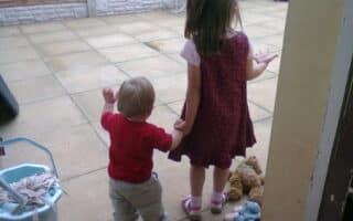 A young girl in a pink and purple dress holds hands with a toddler in a red shirt and light trousers. They stand on a tiled patio near a mop bucket, facing away. Toys, including a teddy bear and a doll, are scattered on the ground beside them.