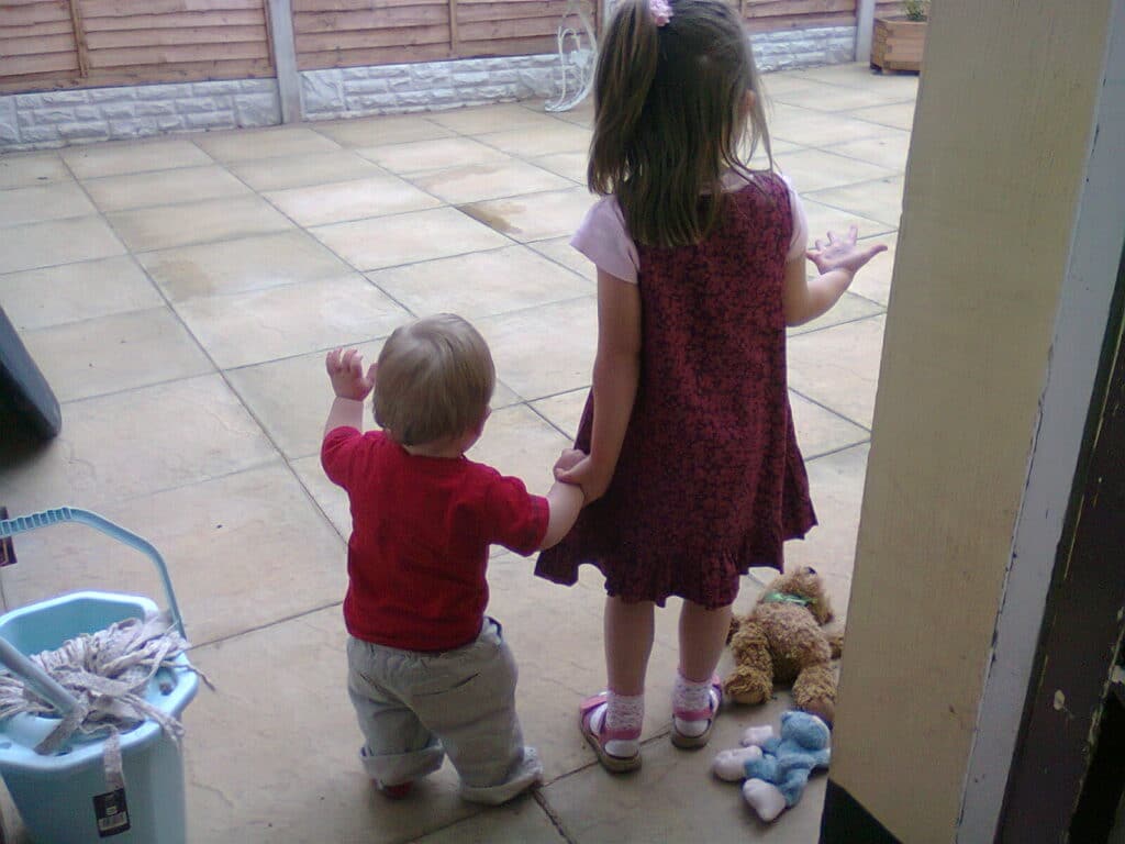 A young girl in a pink dress holds hands with a toddler in a red shirt. They stand on a patio facing away from the camera. To their right, soft toys lie on the ground; to the left, there’s a blue bucket with a mop inside. A wooden fence is in the background.