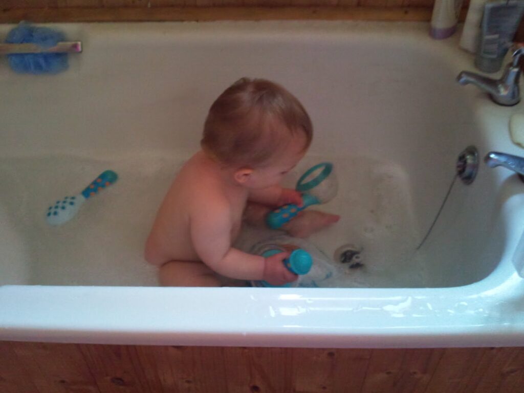 A young child sits in a bath filled with shallow water, holding blue and white bath toys. The child faces sideways, and more toys float nearby. The bath is white with a silver tap and wooden panelling on the outside. Toiletries are visible on the bath rim.
