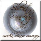 A close-up photo of a camera lens with elegant, cursive text overlay reading: "Silent Sunday, at the mocha beanie mummy." The image has a soft, light background and a faint border, giving it a gentle, artistic effect.