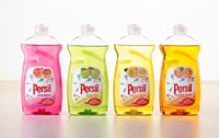 Four bottles of Persil washing-up liquid stand side by side. The liquids are coloured pink, green, yellow, and orange, each with a fruit-themed label. The bottles are clear with white caps, placed on a light surface against a plain background.