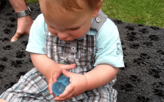 A toddler with blond hair, wearing a blue T-shirt and checked dungarees, sits on a dark blanket outdoors. The child is looking down, holding a blue object in both hands. An adult’s arm is partially visible on the left. The background features green grass.