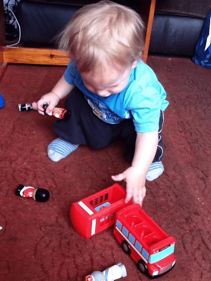 A toddler with blonde hair, wearing a blue shirt and striped socks, kneels on a brown carpet. The child plays with a red toy bus and toy figurines, focusing intently as they touch the bus. Other toys are scattered nearby.