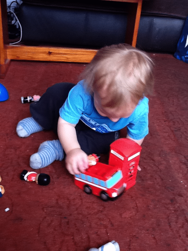 A toddler with fair hair, wearing a blue shirt and striped socks, sits on a red carpet playing with a red toy double-decker bus and small toy figures. The child is focused on placing a figure inside the bus. Furniture and toys are visible in the background.