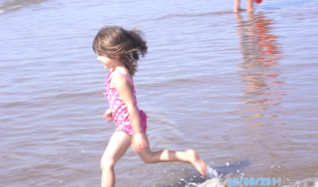 A young girl in a pink swimming costume runs through shallow water at the beach, splashing as she moves. In the background, an adult stands partially visible, holding a red flotation ring. The date "22/08/2011" is visible in the lower right corner.