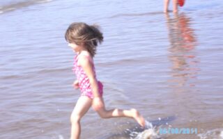 A young girl in a pink swimming costume runs through shallow water at the beach, splashing as she moves. In the background, an adult stands partially visible, holding a red flotation ring. The date "22/08/2011" is visible in the lower right corner.