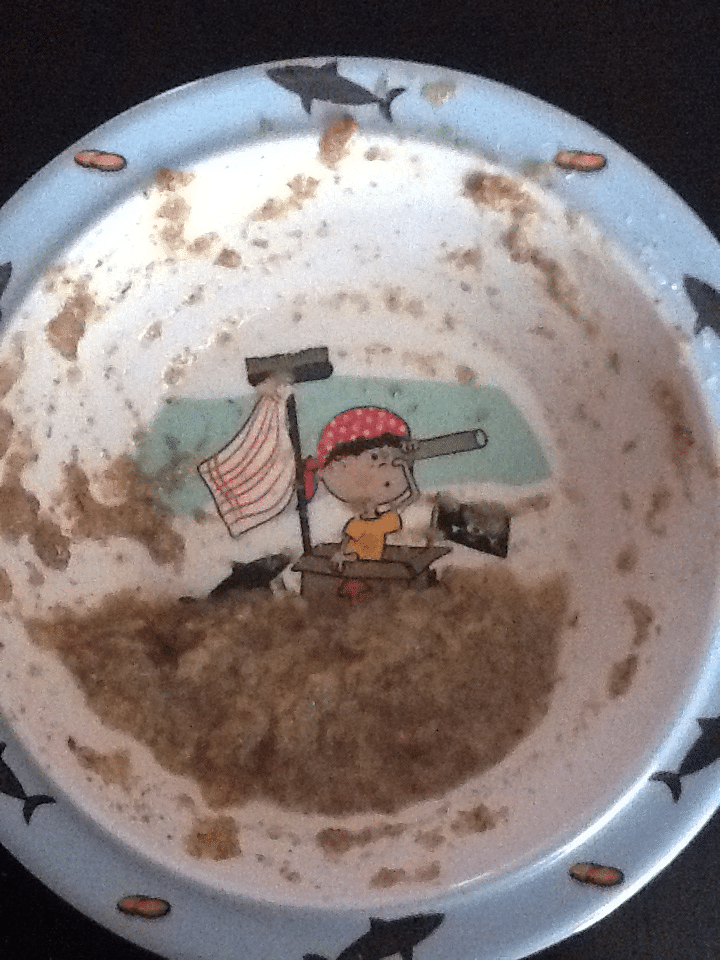 A mostly empty bowl with remnants of brown porridge reveals an illustration of a child in a red polka-dot bandana using a telescope on a small boat with a sail. The bowl's rim is decorated with images of sharks.