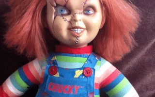 A Chucky doll with messy ginger hair, blue eyes, a sinister grin, and facial scars. It wears a striped long-sleeved shirt and blue dungarees with colourful prints and the name “CHUCKY” in red, sitting against a dark cushion.