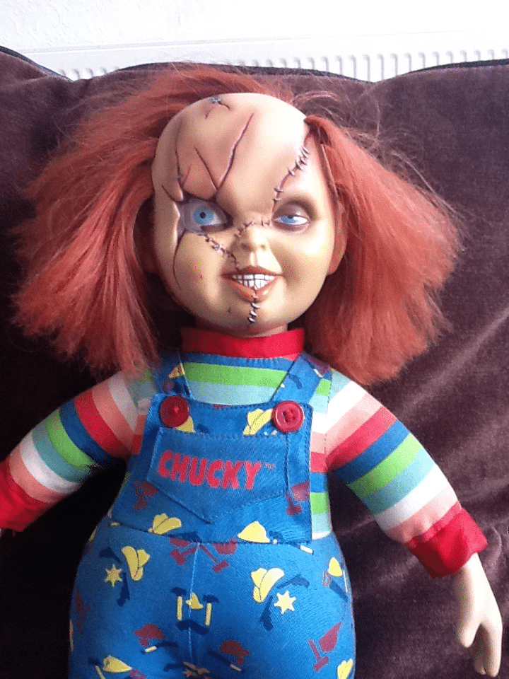 A Chucky doll with messy ginger hair, blue eyes, a sinister grin, and facial scars. It wears a striped long-sleeved shirt and blue dungarees with colourful prints and the name “CHUCKY” in red, sitting against a dark cushion.
