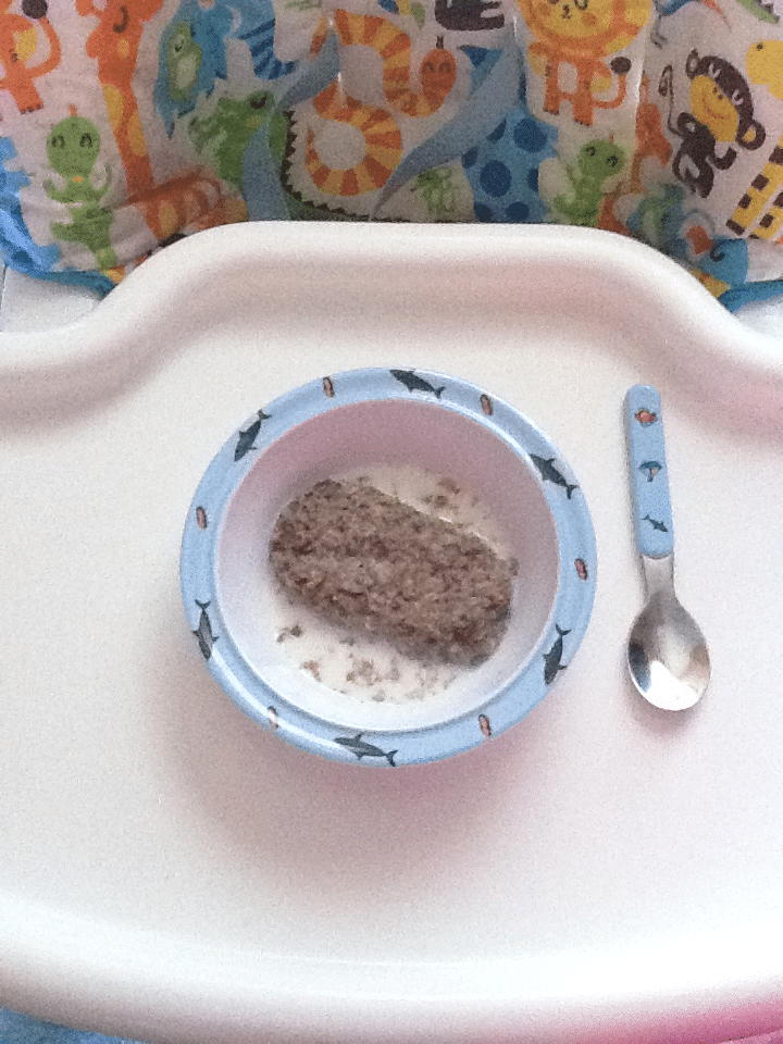 A blue bowl with a single Weetabix biscuit in milk sits on a white tray. A matching blue spoon with animal designs lies to the right. The background shows a colourful fabric with images of animals like lions, giraffes, and crocodiles.