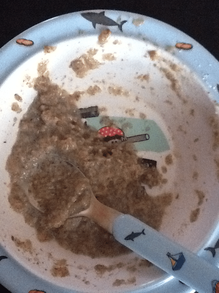 A child’s plate with cartoon sharks and a little food left, possibly porridge or oat porridge. A matching spoon with a shark handle rests in the bowl, surrounded by light brown, lumpy food residue spread unevenly across the plate.
