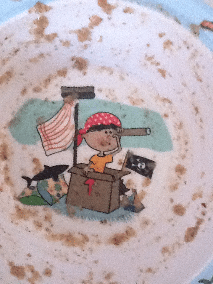 A plate with food crumbs reveals a cartoon of a child wearing a red polka-dot bandana, looking through a telescope from a cardboard box pirate ship. A pirate flag, striped sail, shark, and treasure chest are in the background.