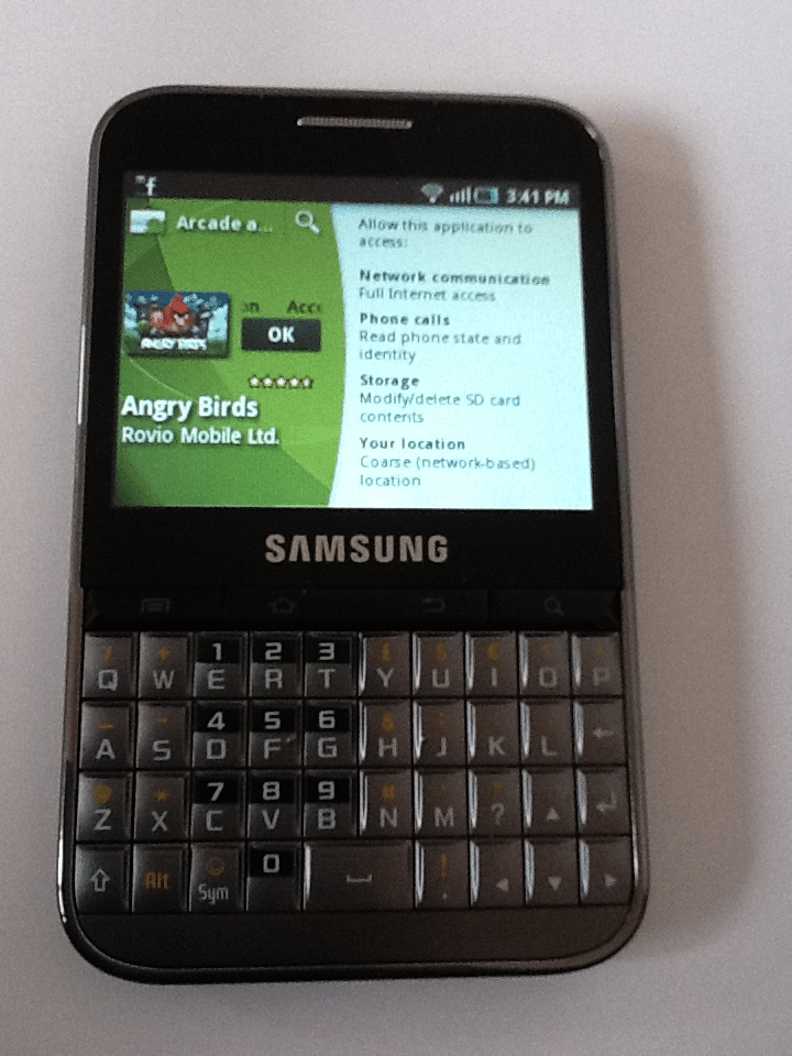 A Samsung smartphone with a physical QWERTY keyboard is shown. The screen displays the Angry Birds app permissions in the Google Play Store, with the app icon and some permission details visible. The phone is black and sits on a white surface.