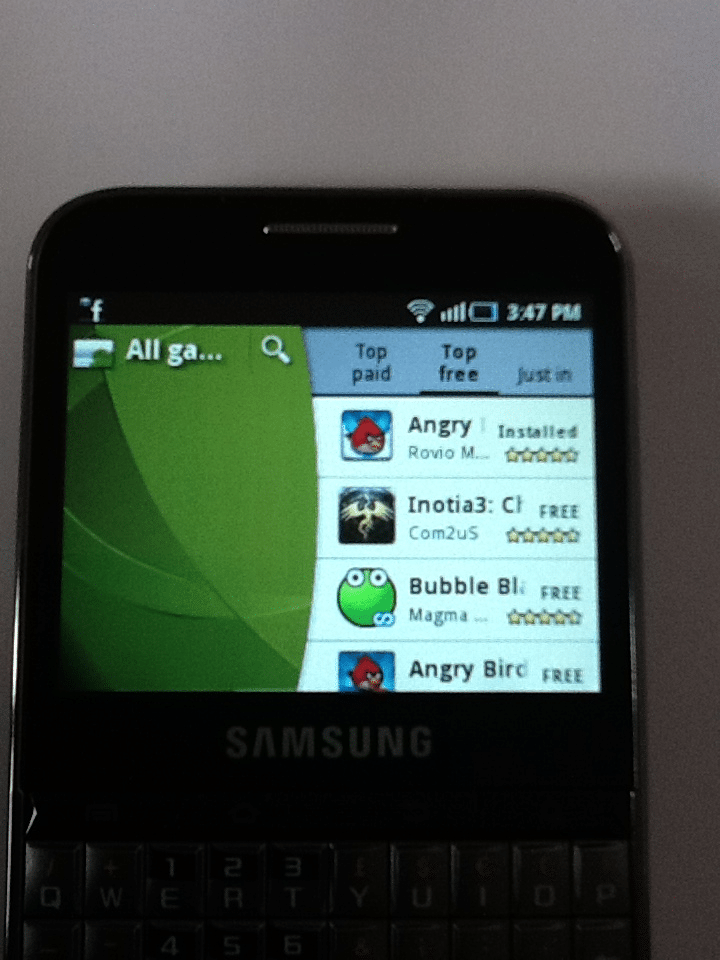 A Samsung smartphone displays a screen showing a list of mobile games, including "Angry Birds," "Inotia3: Children of Carnia," and "Bubble Blast 2," with app icons, ratings, installation status, and tabs for "Top paid," "Top free," and "Just in.