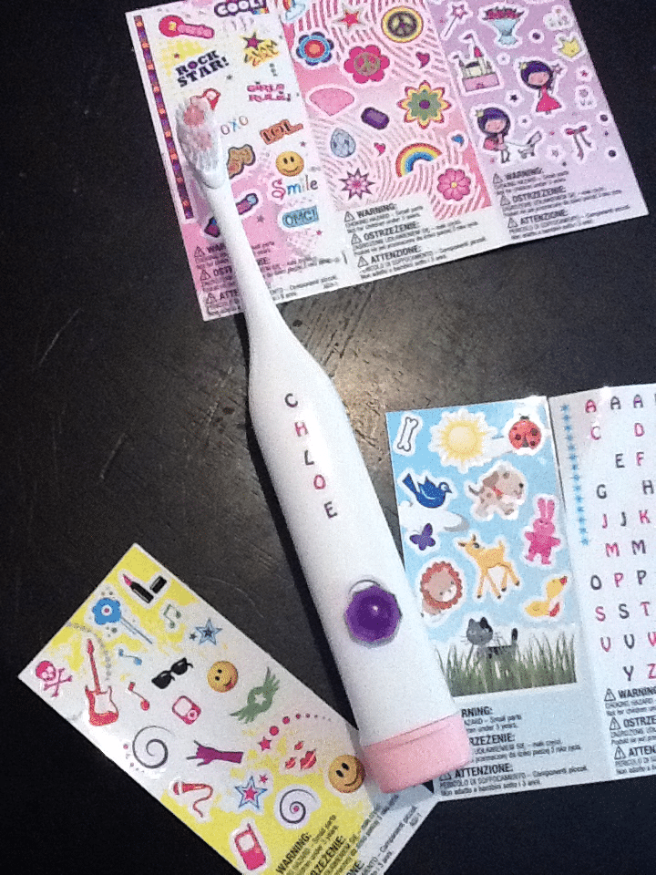 A white electric toothbrush labelled "CHLOE" is placed on a dark surface, surrounded by colourful sticker sheets with letters, emojis, stars, flowers, musical notes, and various fun designs intended for decorating items.