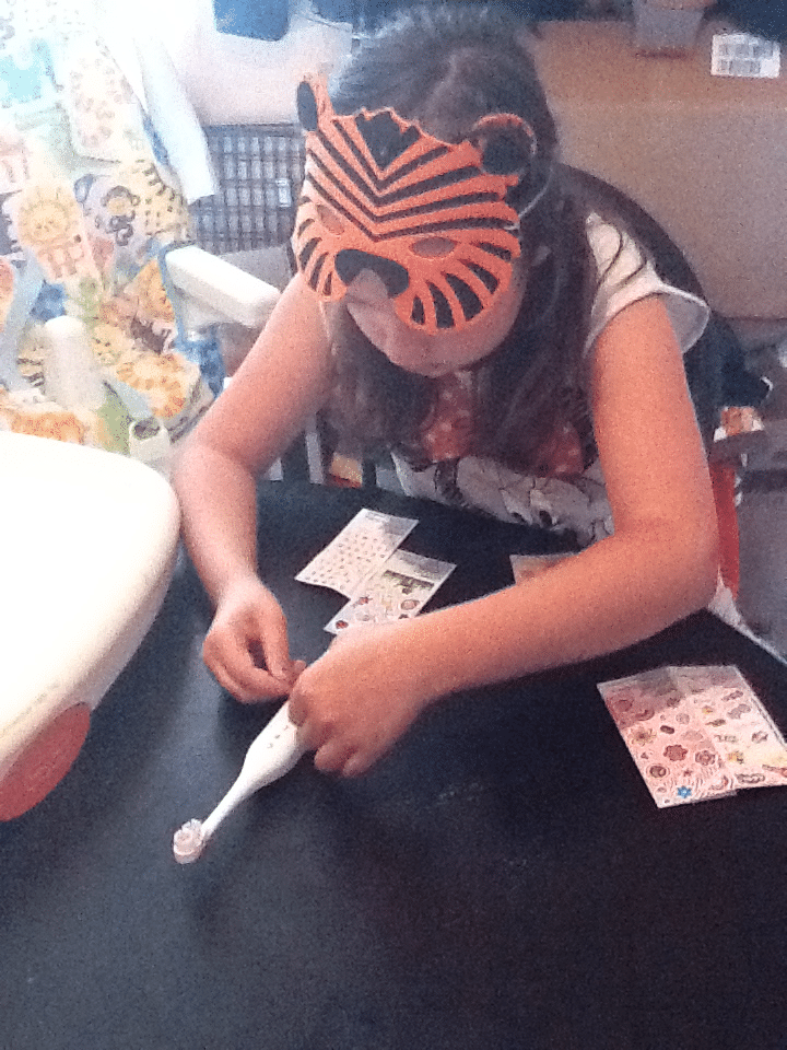 A young girl wearing an orange tiger mask sits at a dark table, decorating a white electric toothbrush with colourful stickers from sticker sheets. A patterned high chair is visible in the background. The scene appears playful and creative.