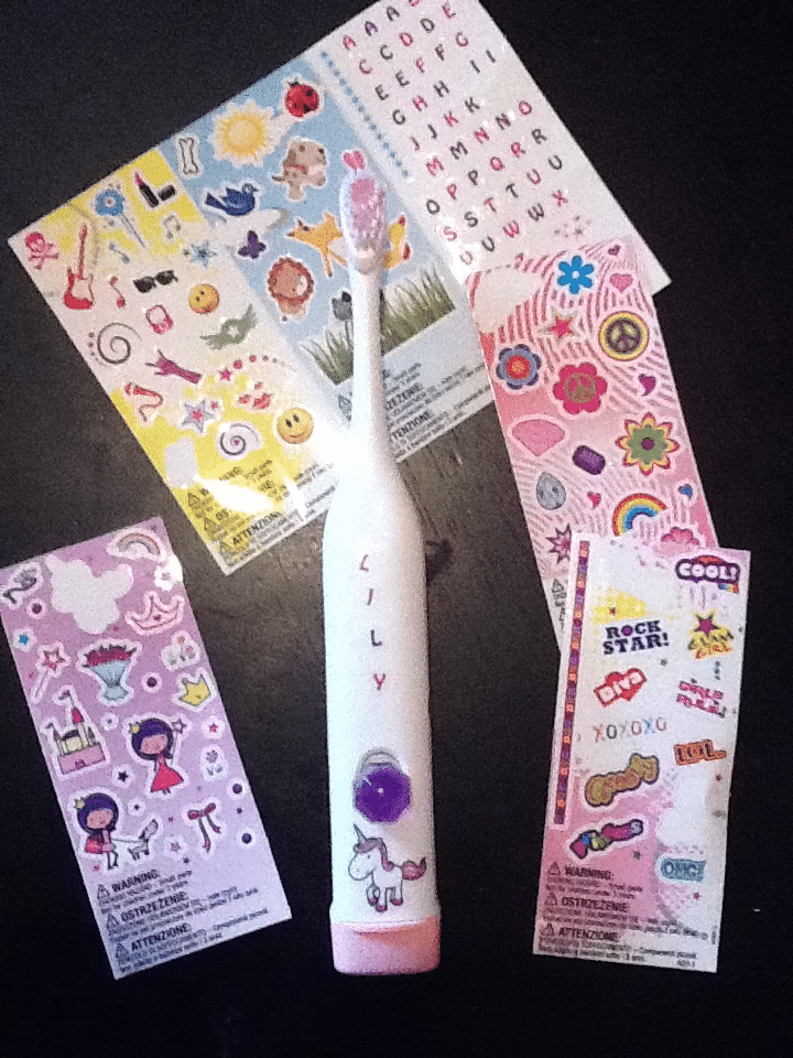An electric toothbrush labelled "LILY" with a unicorn sticker is surrounded by colourful sticker sheets featuring stars, hearts, animals, smiley faces, and cartoon characters on a dark surface.