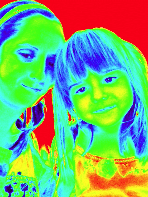 A false-colour thermal-like image shows a woman and a young girl close together, smiling. Their faces appear in blue and green hues, hair in blue and yellow tones, while the background is bright red, creating a vivid, high-contrast effect.